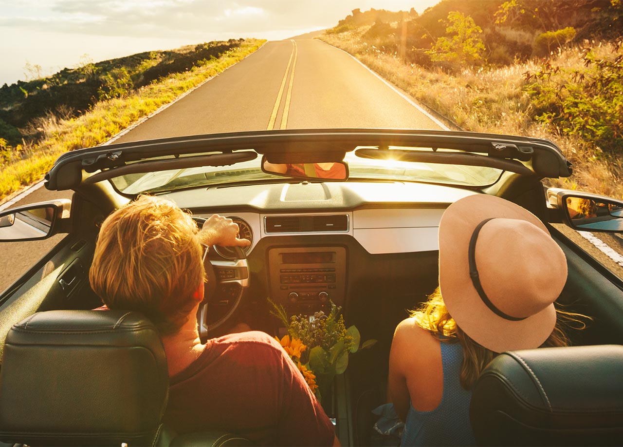 Two people are driving in a convertible car on a sunny road surrounded by grassy hills Auto Insurance - proinsurancenc.com